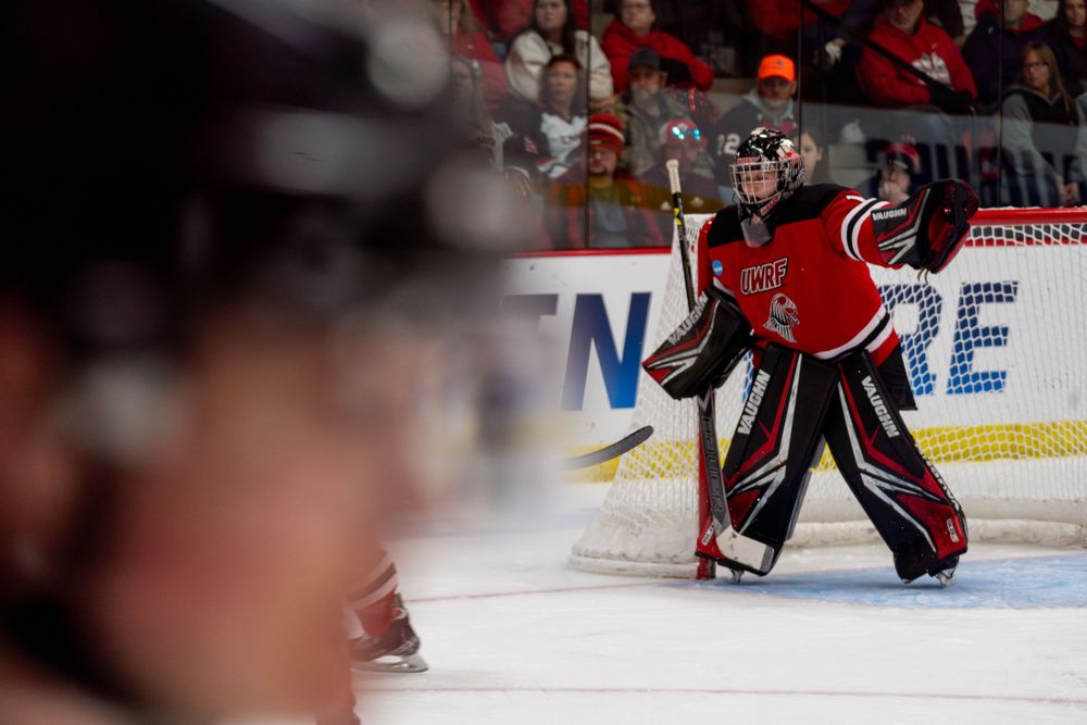 Hockey goalie in red jersey stands ready in front of the net during an NCAA game.