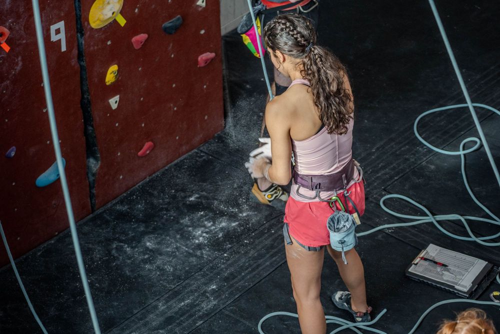 Rock climber in pink tank top and red shorts grips holds on an indoor climbing wall while wearing safety gear.