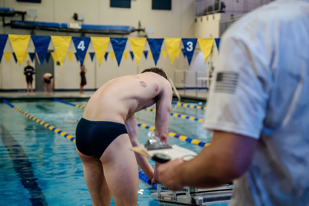 Competitive swimmer on starting block prepares to dive into indoor pool as coach stands nearby with stopwatch.
