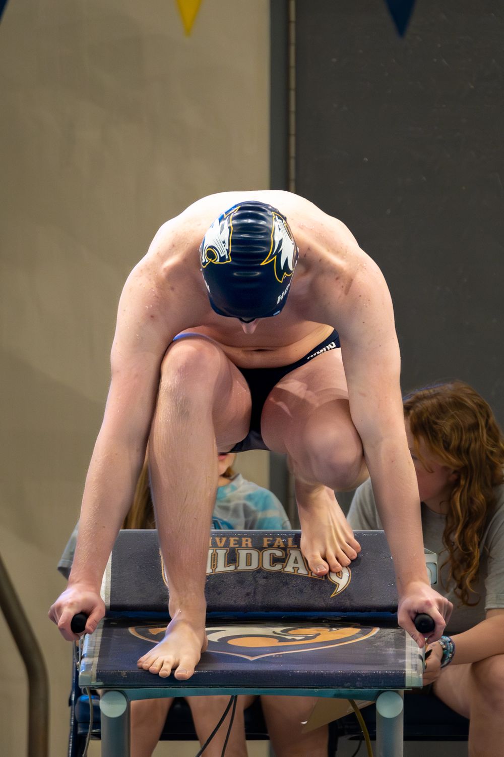 A competitive swimmer in a blue cap crouches at starting position on a diving block before a race.