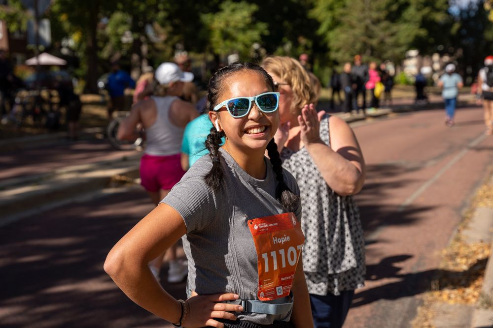 Runner wearing sunglasses and a gray shirt smiles brightly during an outdoor race on a sunny autumn day.