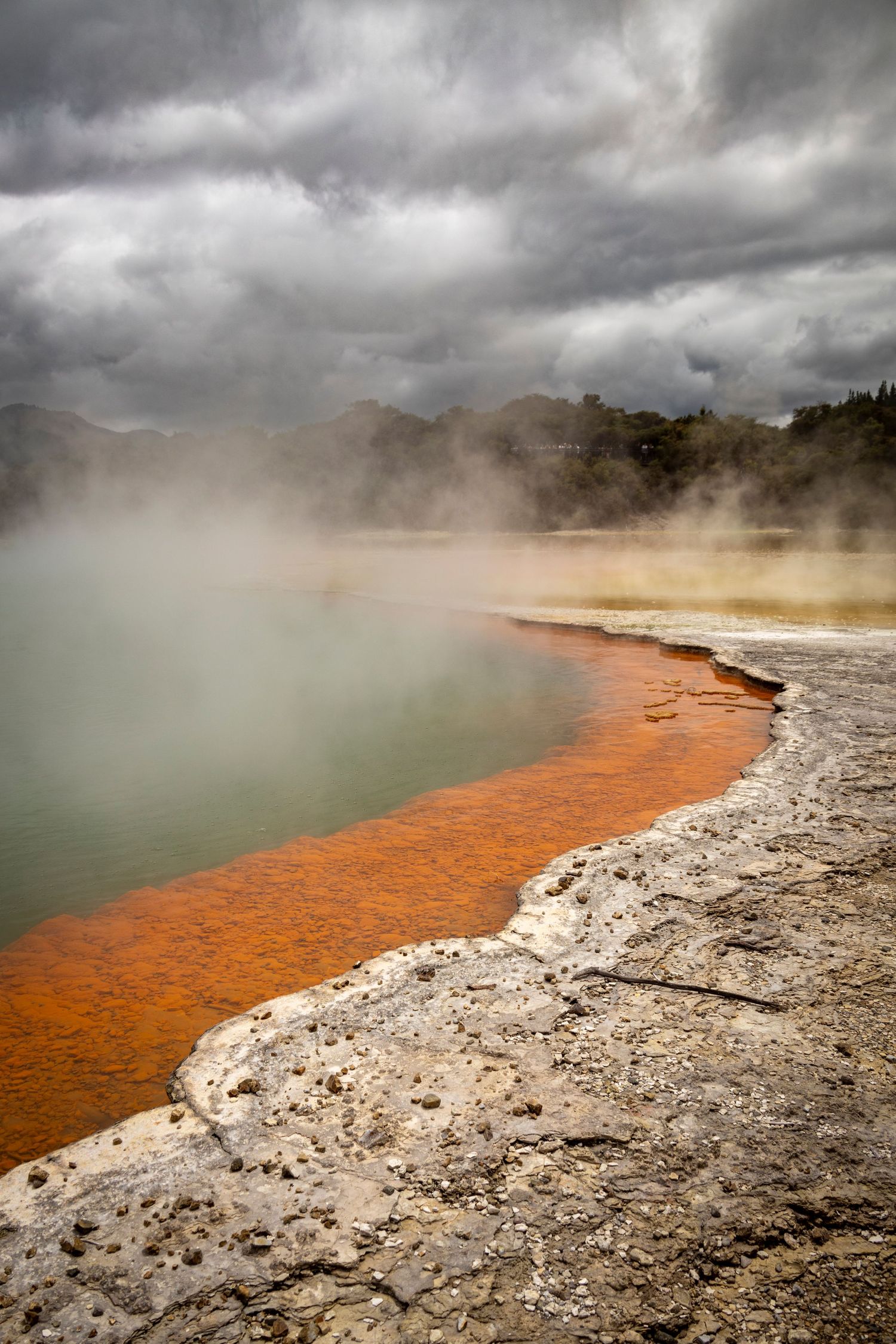 Steaming geothermal pool with orange mineral edge under dramatic clouds, surrounded by rocky terrain.