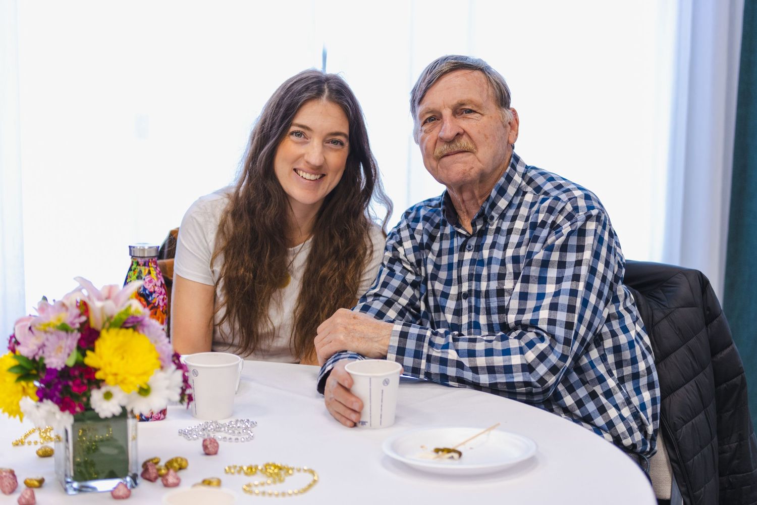 Two people sit at a table set for afternoon tea with flowers and pastries in a bright interior setting.