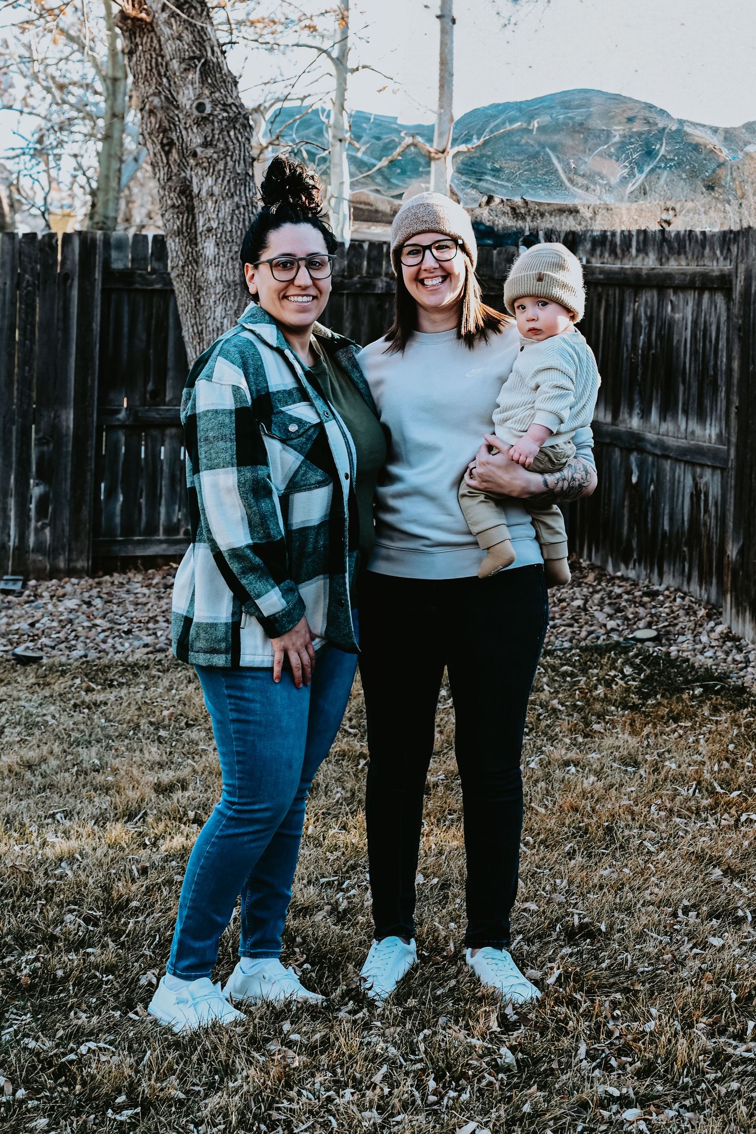 Two women wearing glasses smile outdoors, one holding a baby, with mountains and a wooden fence behind them.