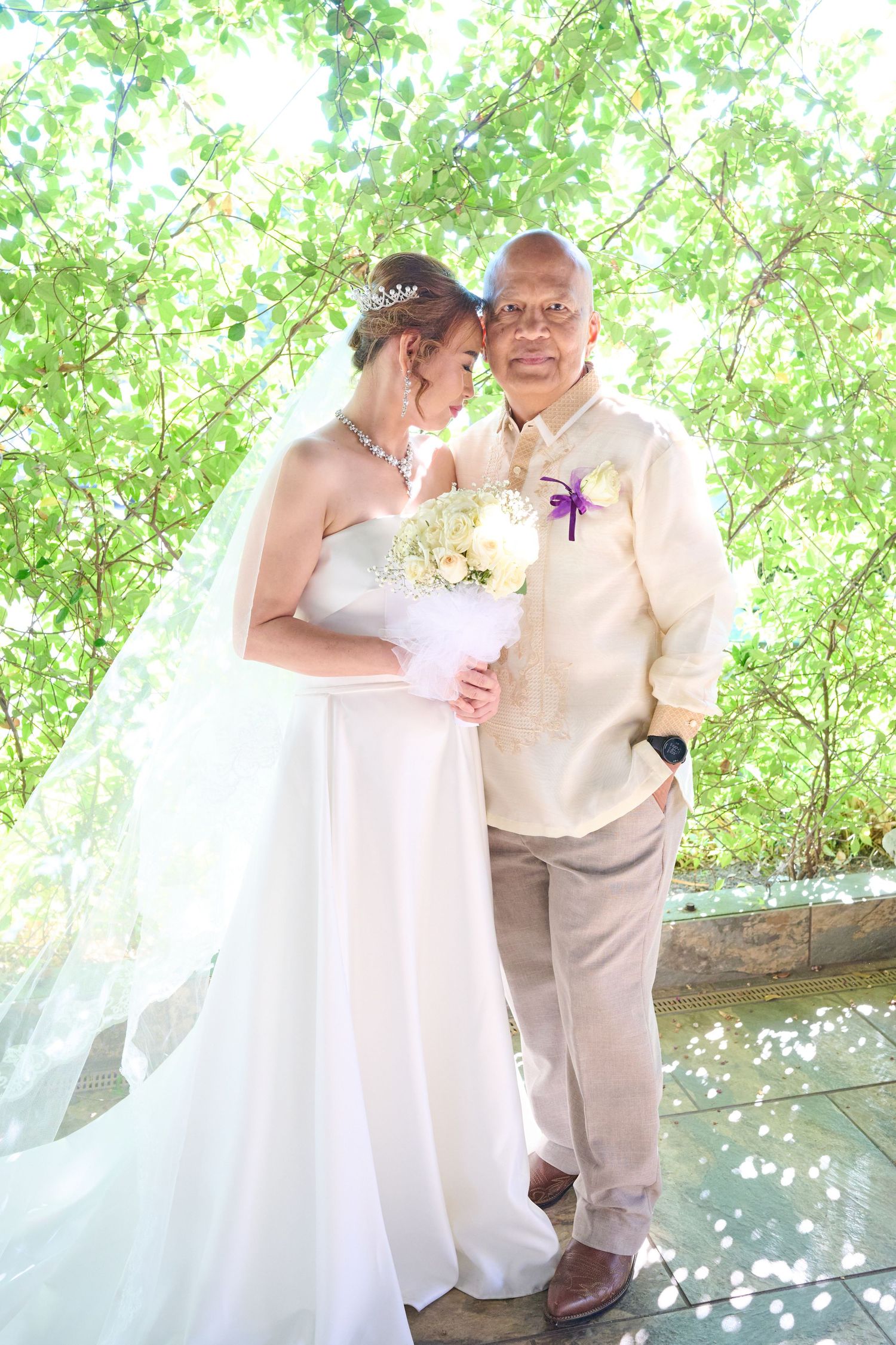 Wedding couple poses in front of religious mural with Virgin Mary figure, bride in white dress and groom in beige suit.