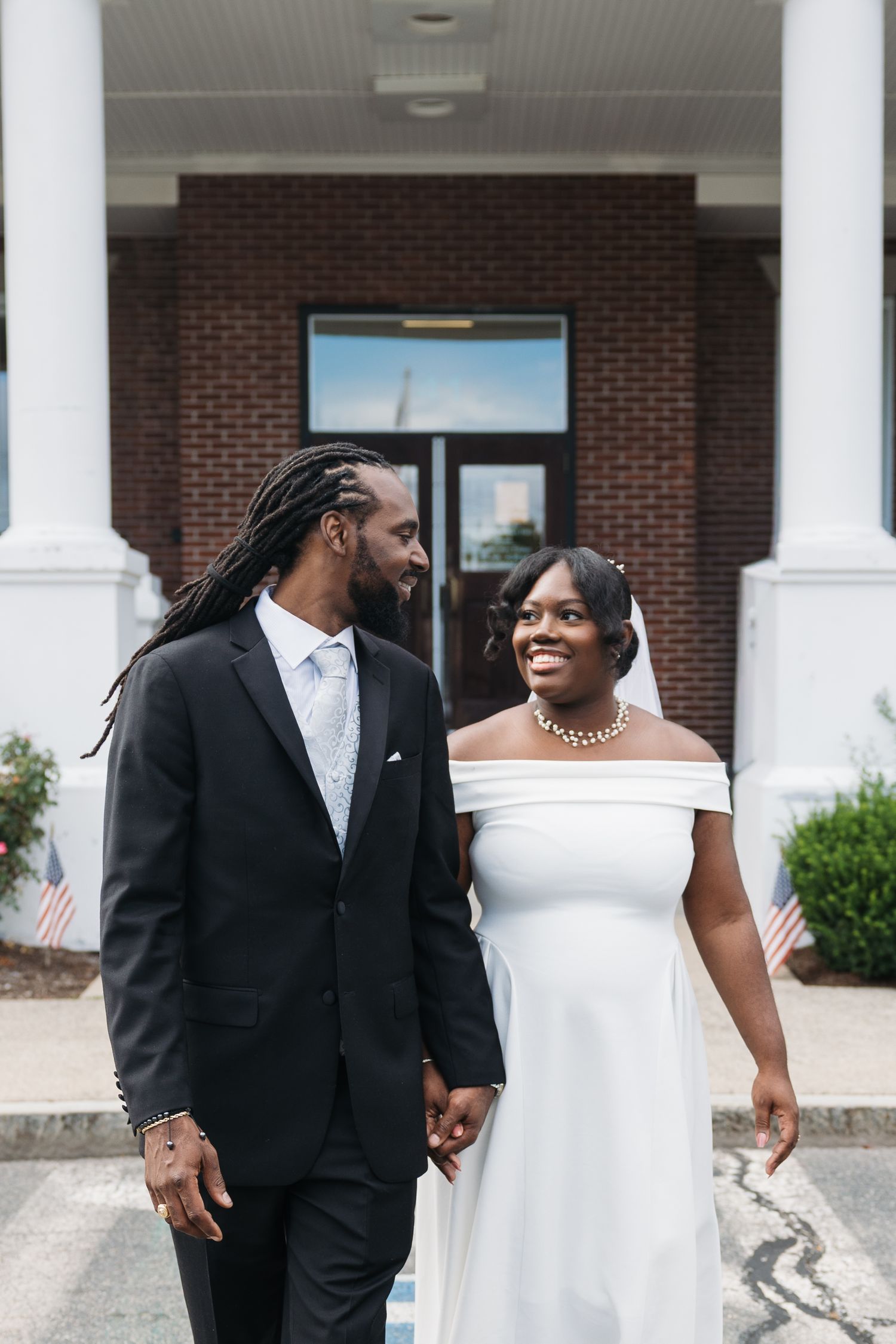 A couple in formal wedding attire share a tender moment between white columns at a brick building entrance.