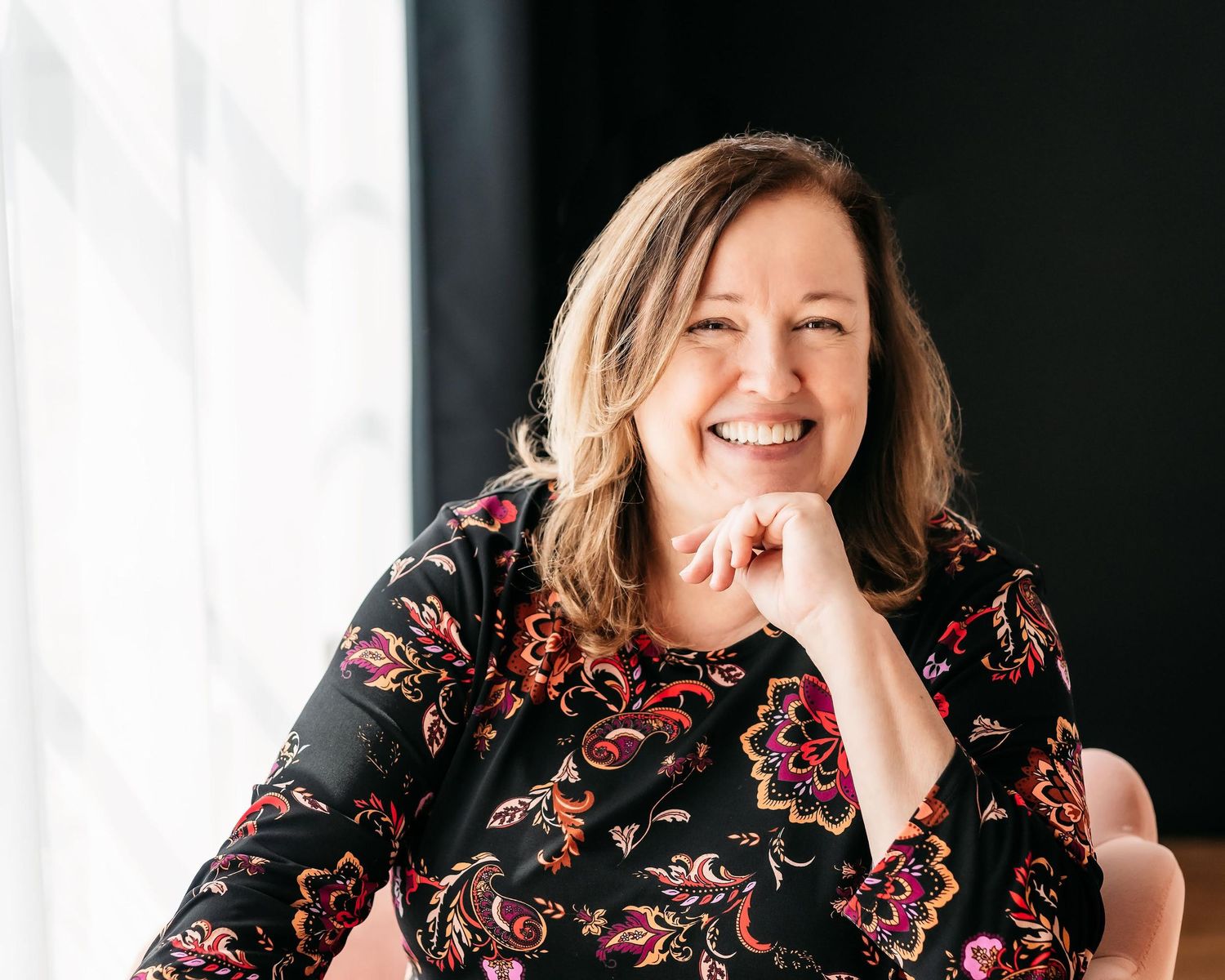 Professional portrait of someone in a floral print top leaning on their hand while smiling against a light background.