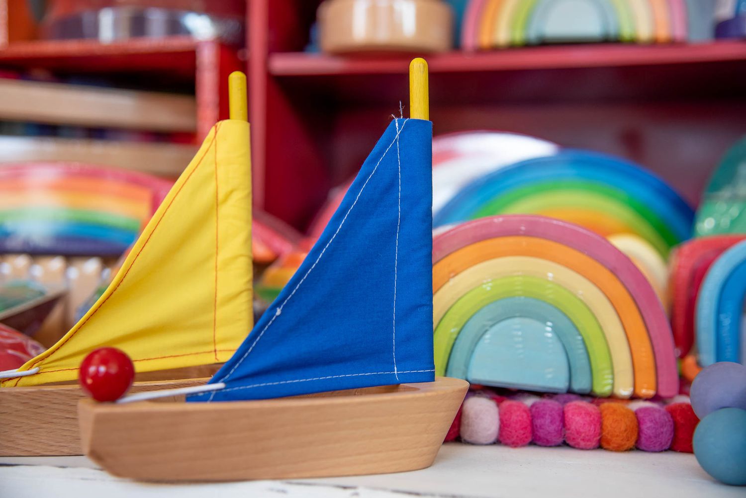 Colorful wooden toy sailboats with yellow and blue sails displayed next to rainbow decorations in a playroom setting.