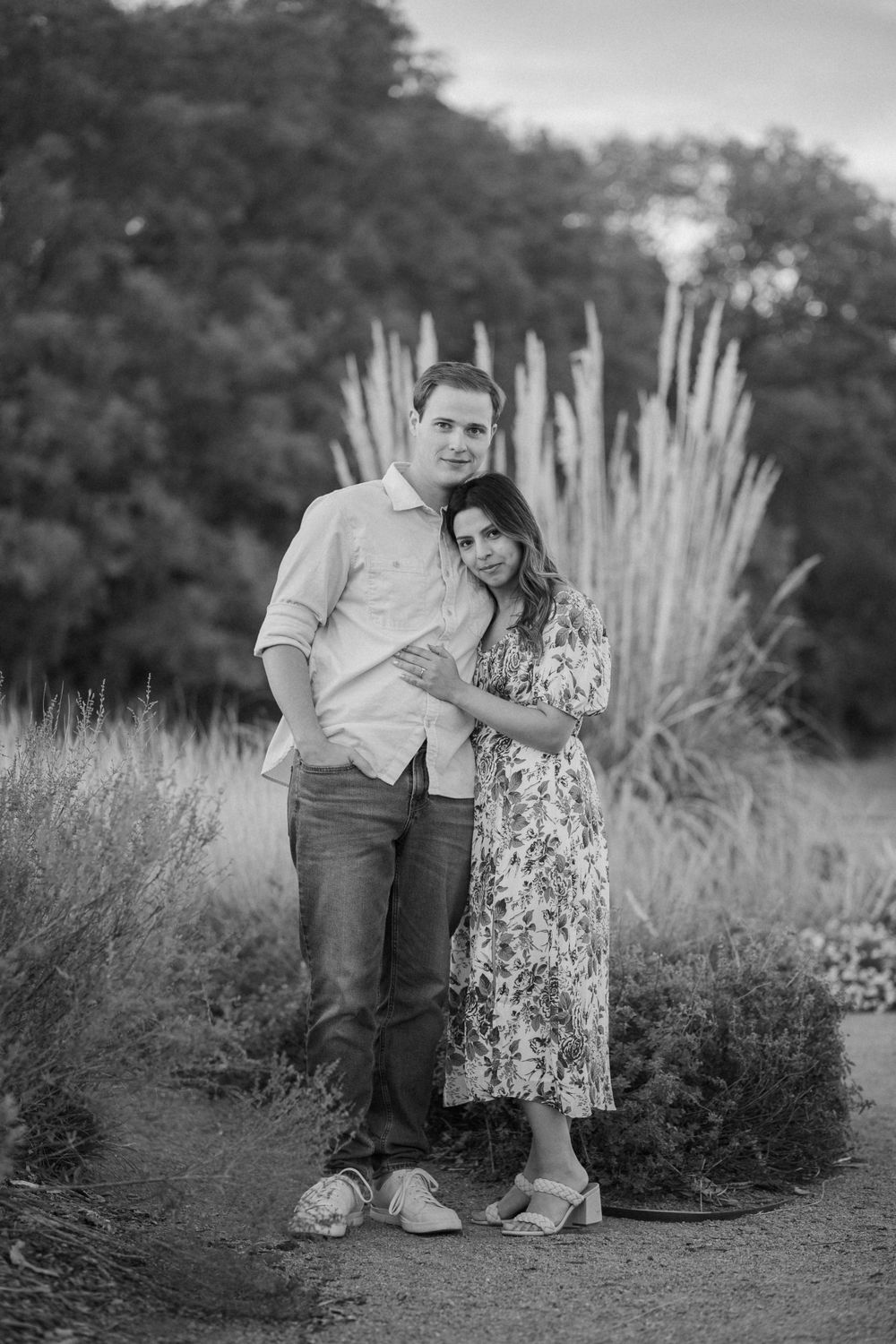 A black and white portrait of a couple standing together on a path surrounded by tall ornamental grasses.