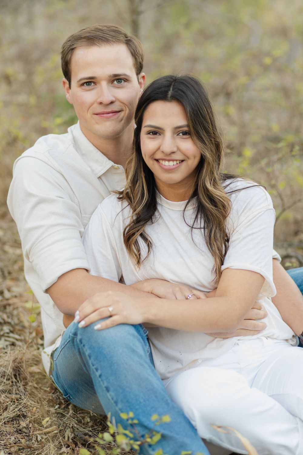 A couple in white shirts sitting together outdoors for an engagement photo.