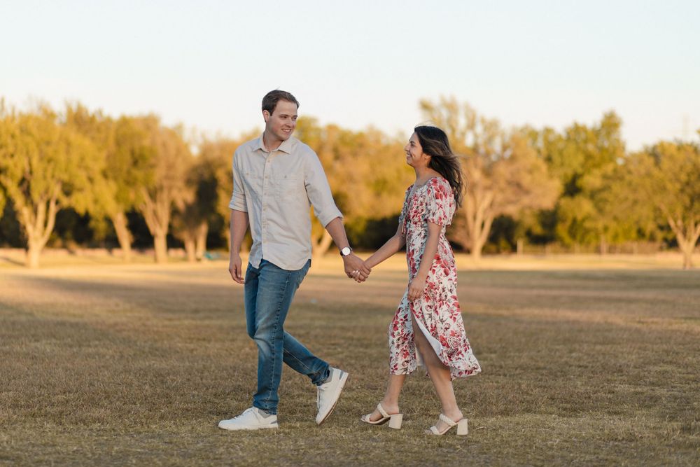 A couple holds hands while walking together in a golden-lit field during sunset.