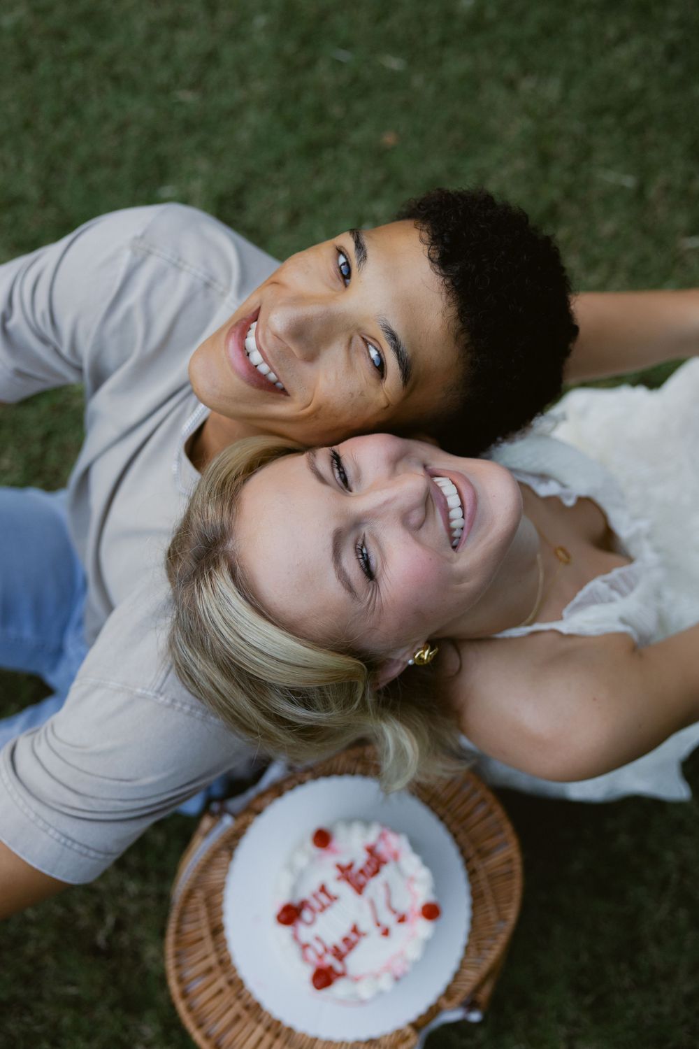 Couple lying on grass sharing a plate of red berries while smiling and enjoying a romantic outdoor moment.