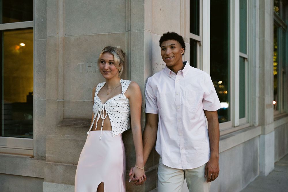 A couple in white outfits walks together while holding hands in front of a stone building exterior.