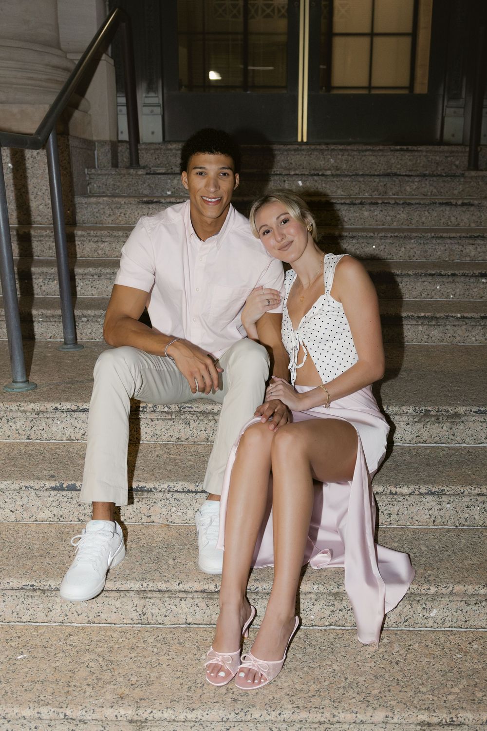 A couple sits casually on concrete steps at night, dressed in white and polka dot outfits.