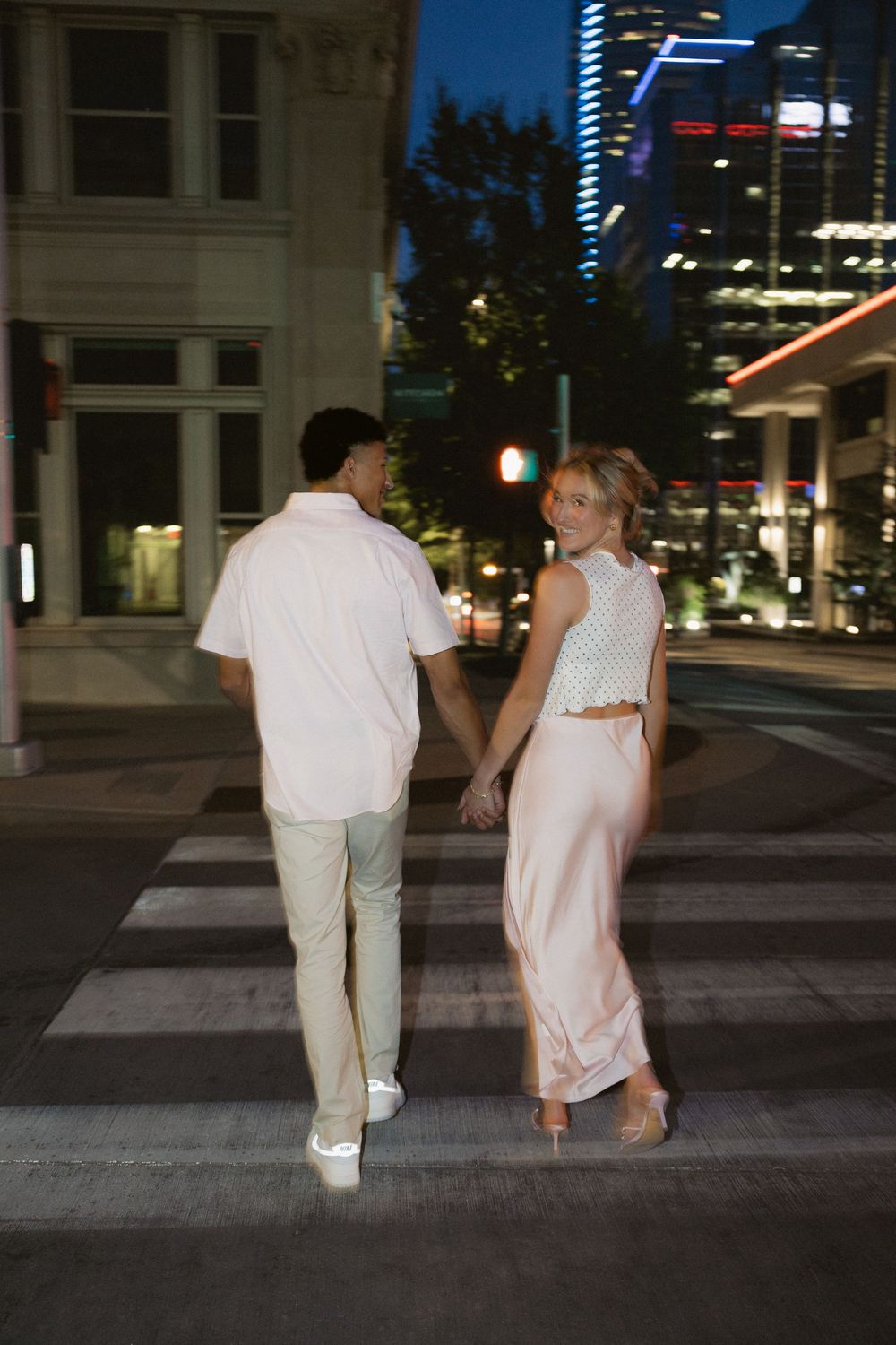 Two people in white outfits walking together at night in an urban setting with illuminated buildings in the background.