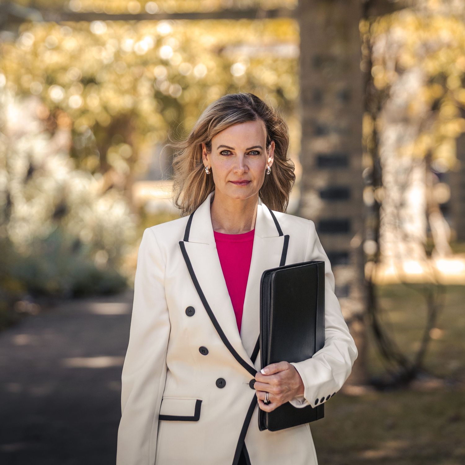 Professional wearing a crisp white blazer with black trim and pink top holds a portfolio folder in an outdoor setting.