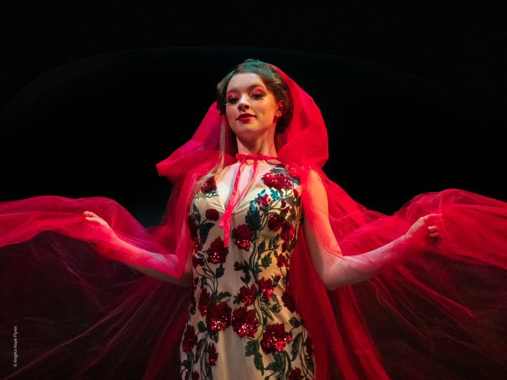 A performer in a floral dress stands dramatically beneath flowing red fabric on stage.