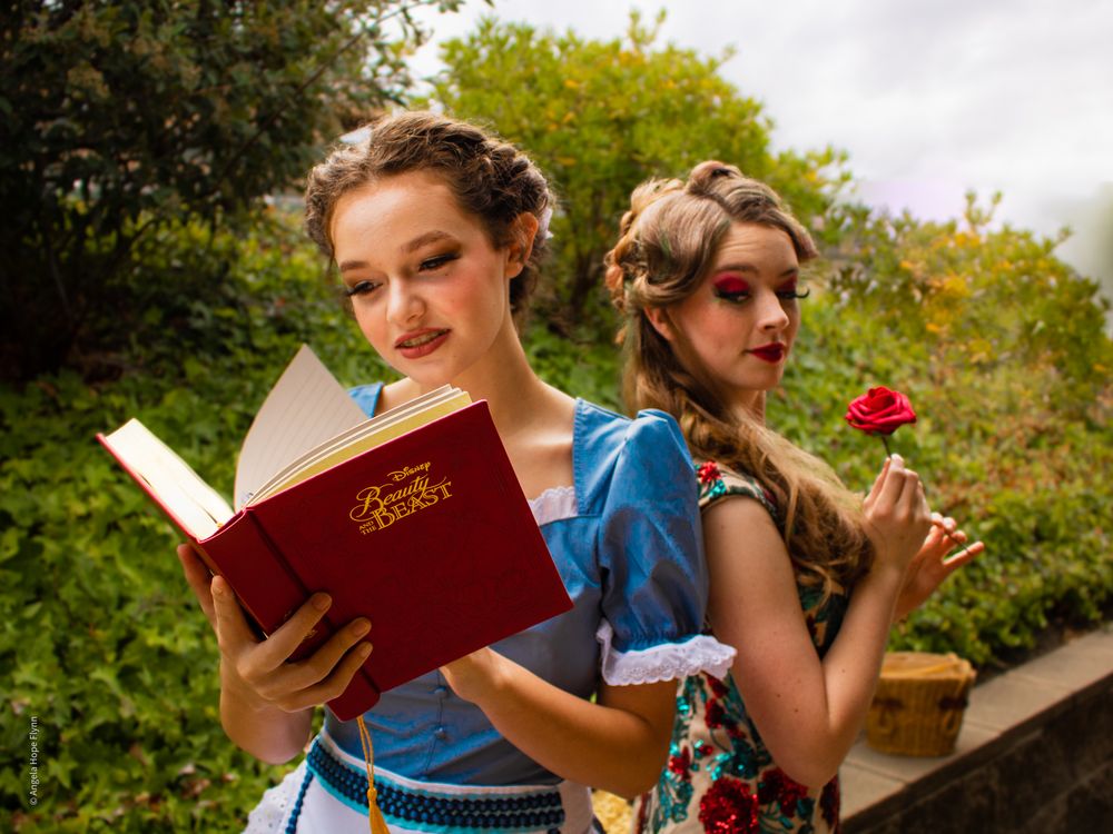 Two performers in blue dresses pose with a book and rose in an outdoor garden setting.