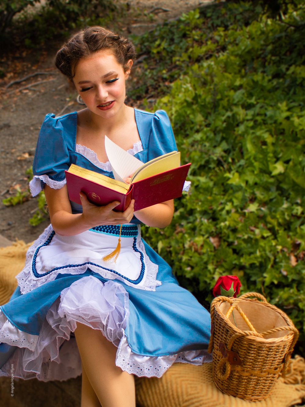 A person in a blue dress with white apron relaxes on a stone wall while reading a book.
