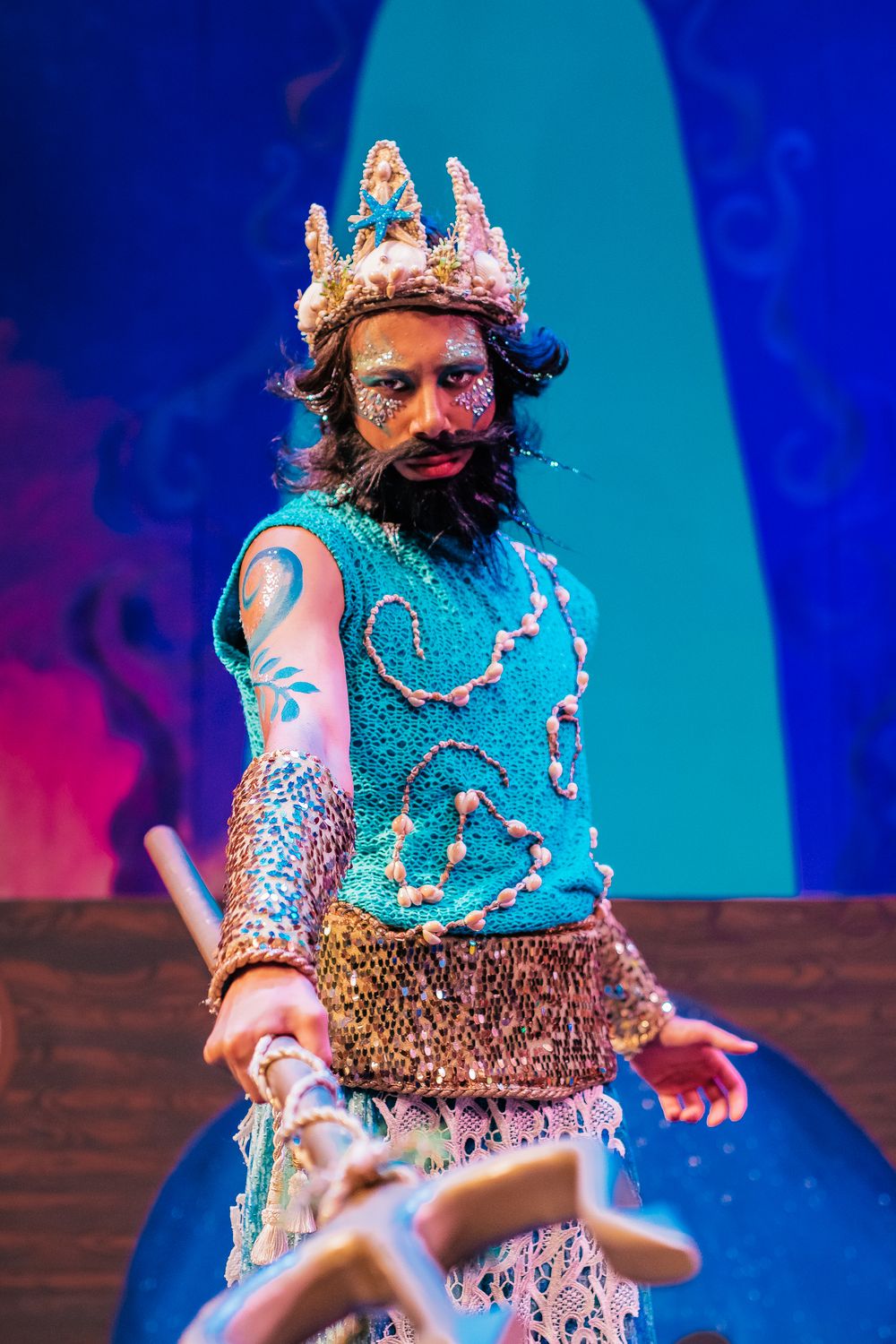 Performer wearing an ornate crown and turquoise costume with metallic details poses dramatically on stage.