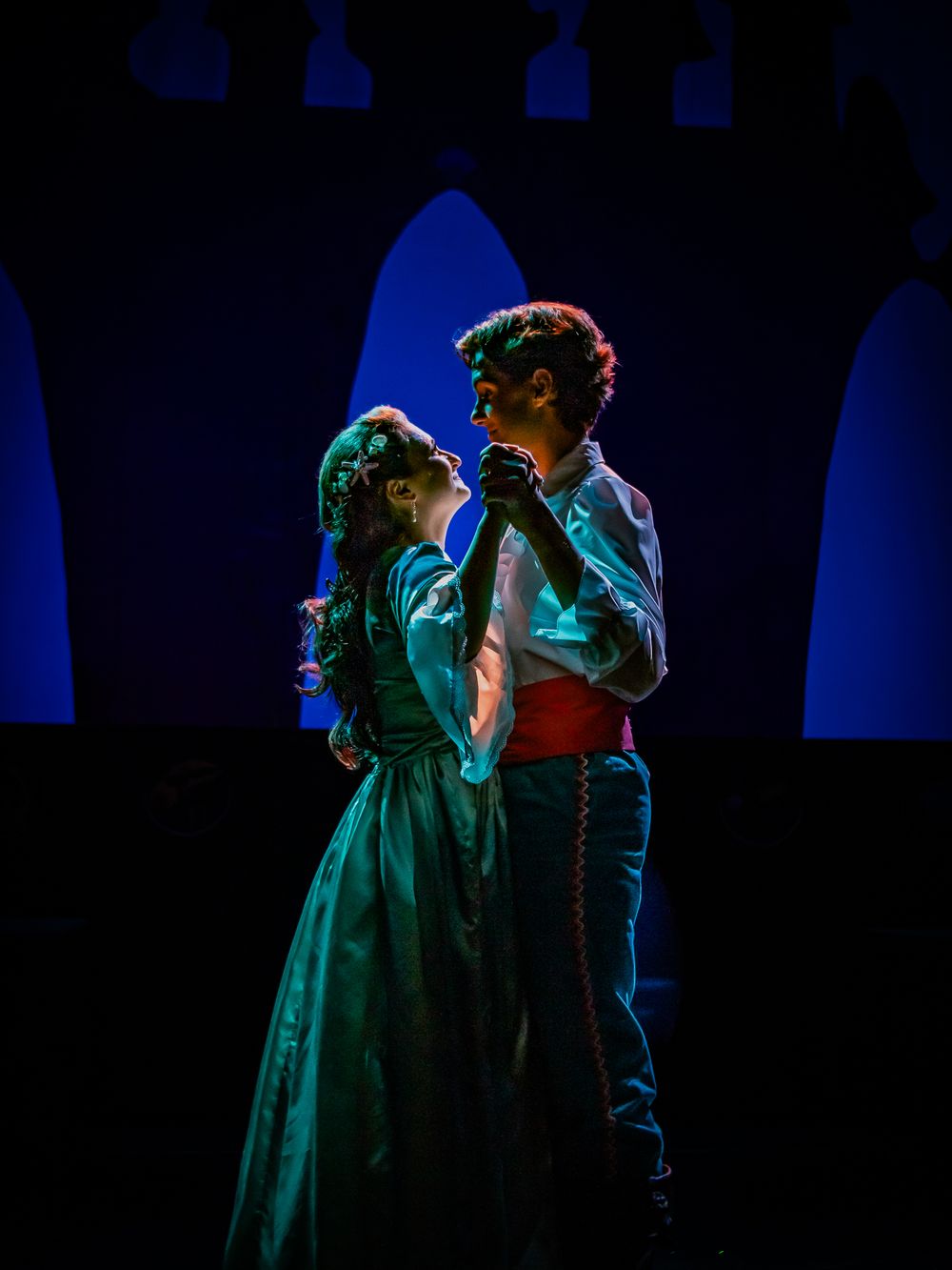 Two performers share an intimate moment in period costumes under teal spotlights.