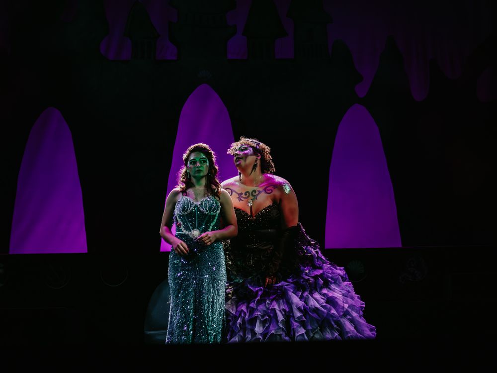 Two performers in glamorous gowns perform on stage with purple archways in the background.