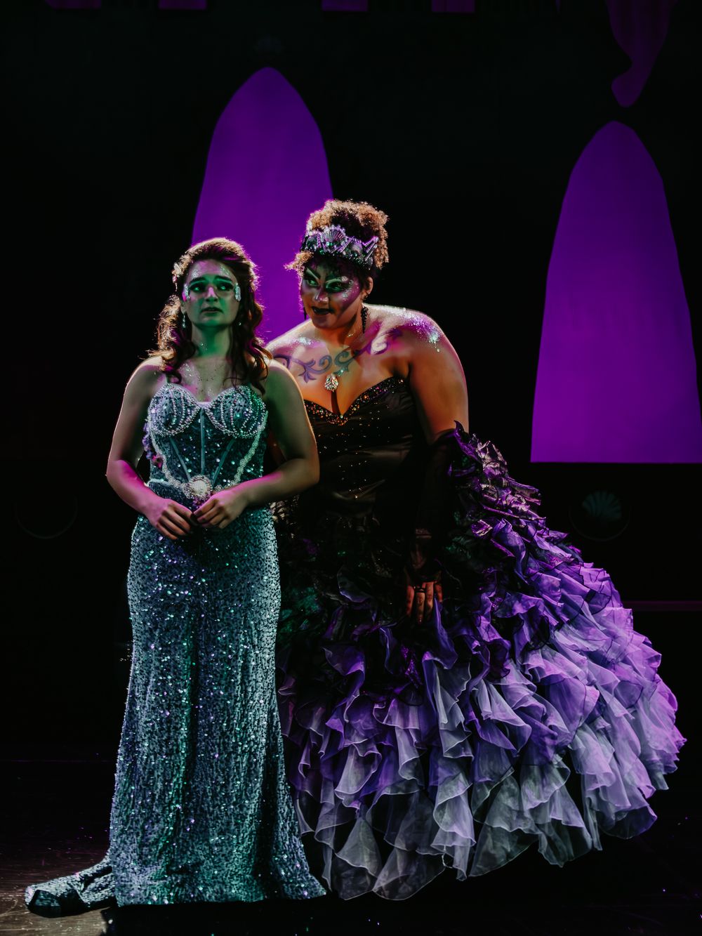 Two stage performers wear elaborate gowns and masks under dramatic purple lighting.