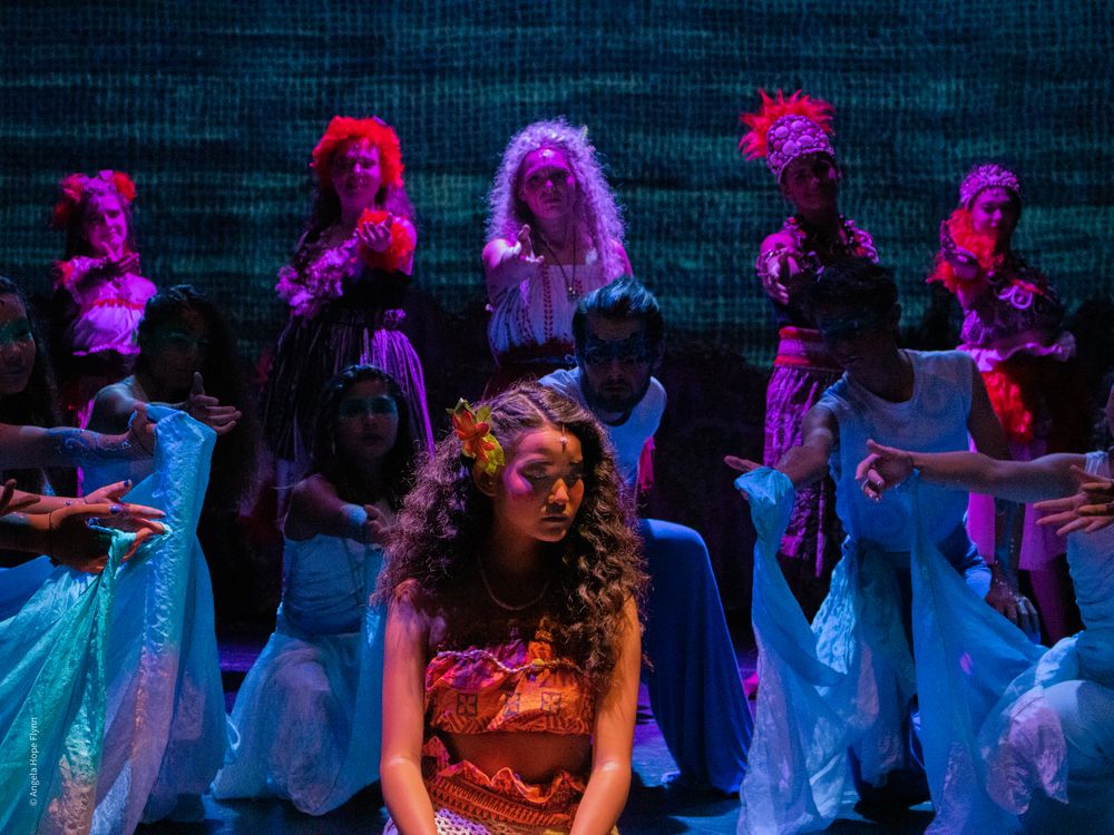 Theater performers in colorful costumes illuminated by vibrant pink and blue stage lighting.