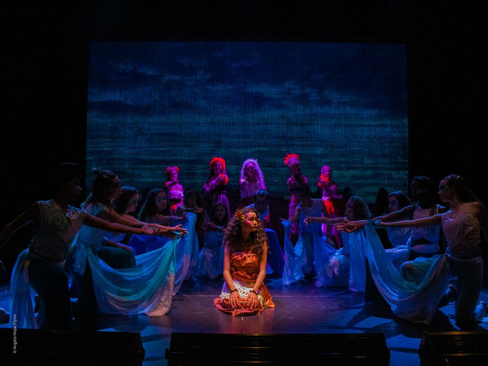 Stage performers in a dramatic theater scene with blue lighting and flowing costumes.