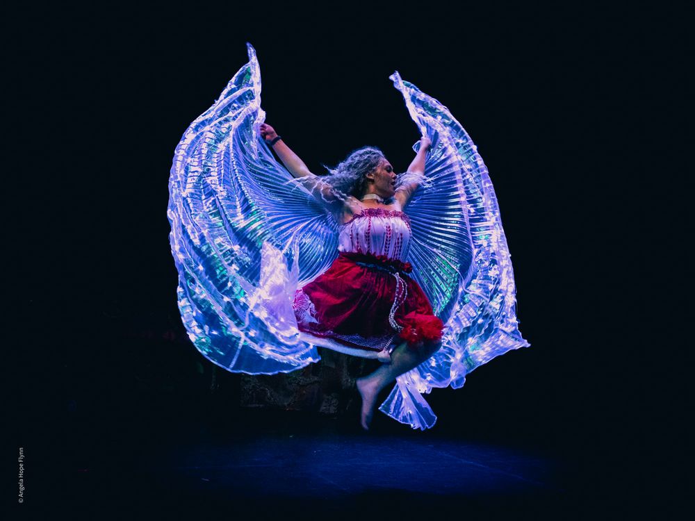 A performer in illuminated wings creates a striking silhouette against a dark background.