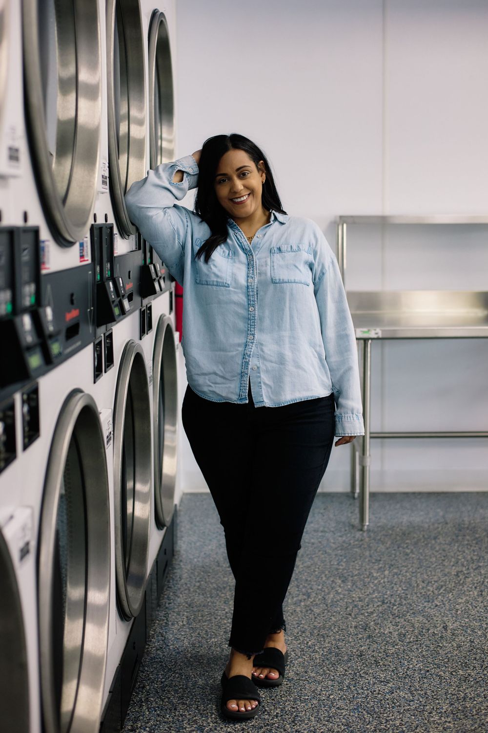 Customer standing in a laundromat between rows of commercial washing machines wearing denim shirt and black pants.