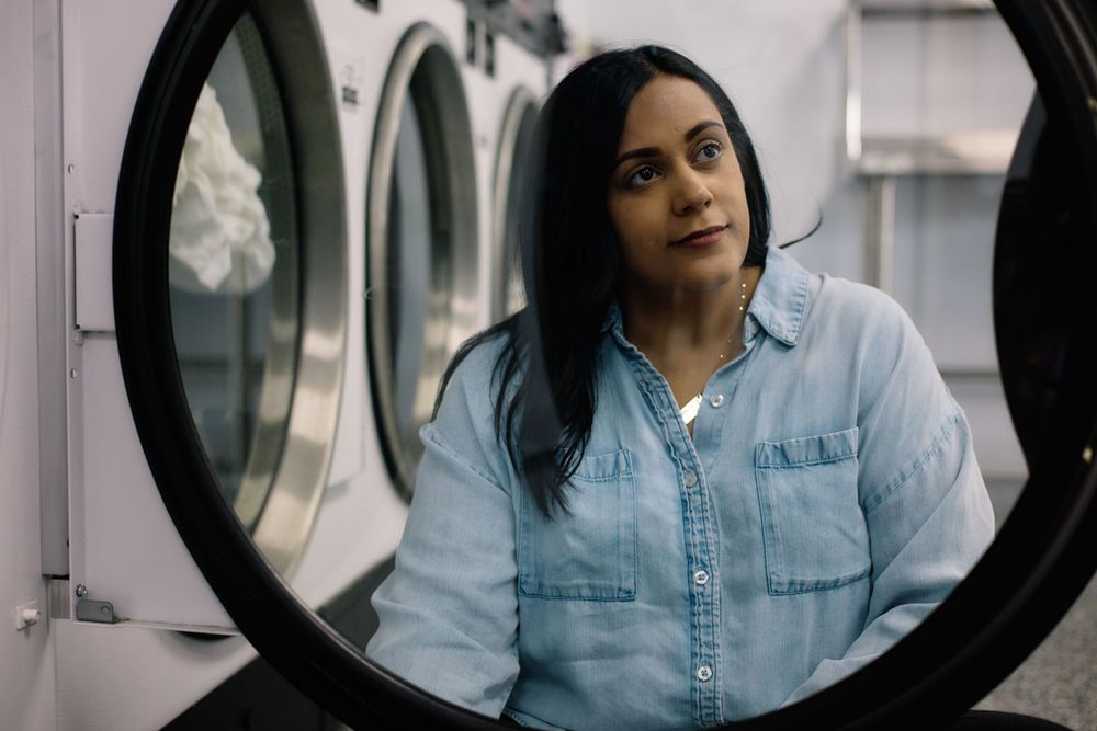 Someone peers through the circular window of a laundromat washing machine while wearing a light blue denim shirt.