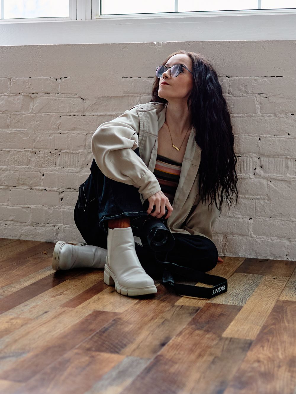 Trendy streetwear outfit featuring white boots, dark pants, cream jacket, and sunglasses on wooden floor against brick wall.