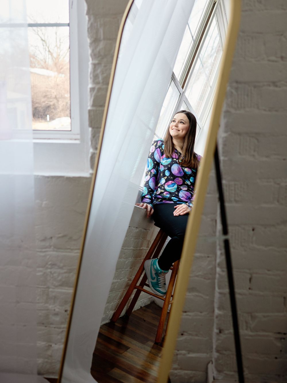 Reflection in mirror shows a casual moment by a bright window, with a floral patterned top and sunlit white walls.