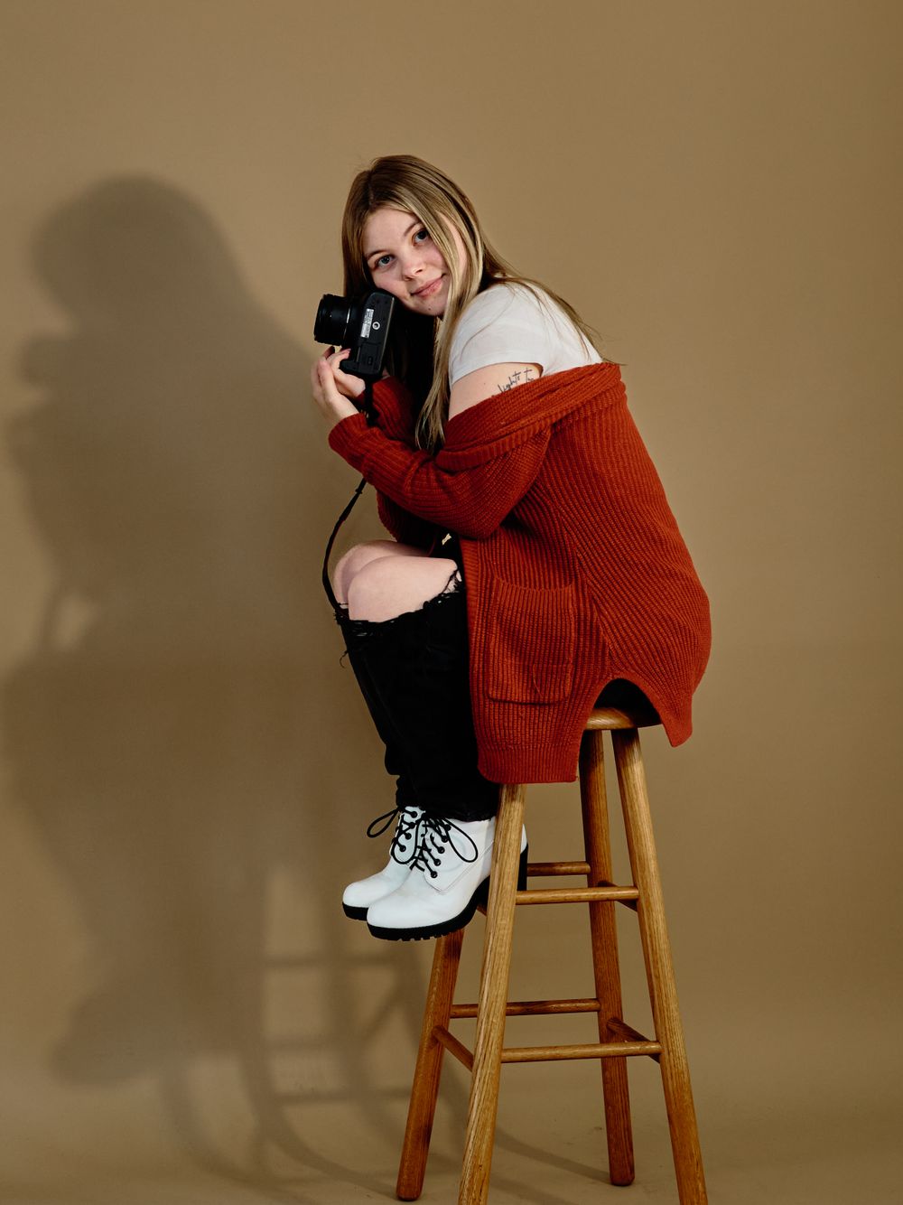 Person in red knit cardigan and white sneakers sits perched on wooden stool against beige backdrop during photoshoot.