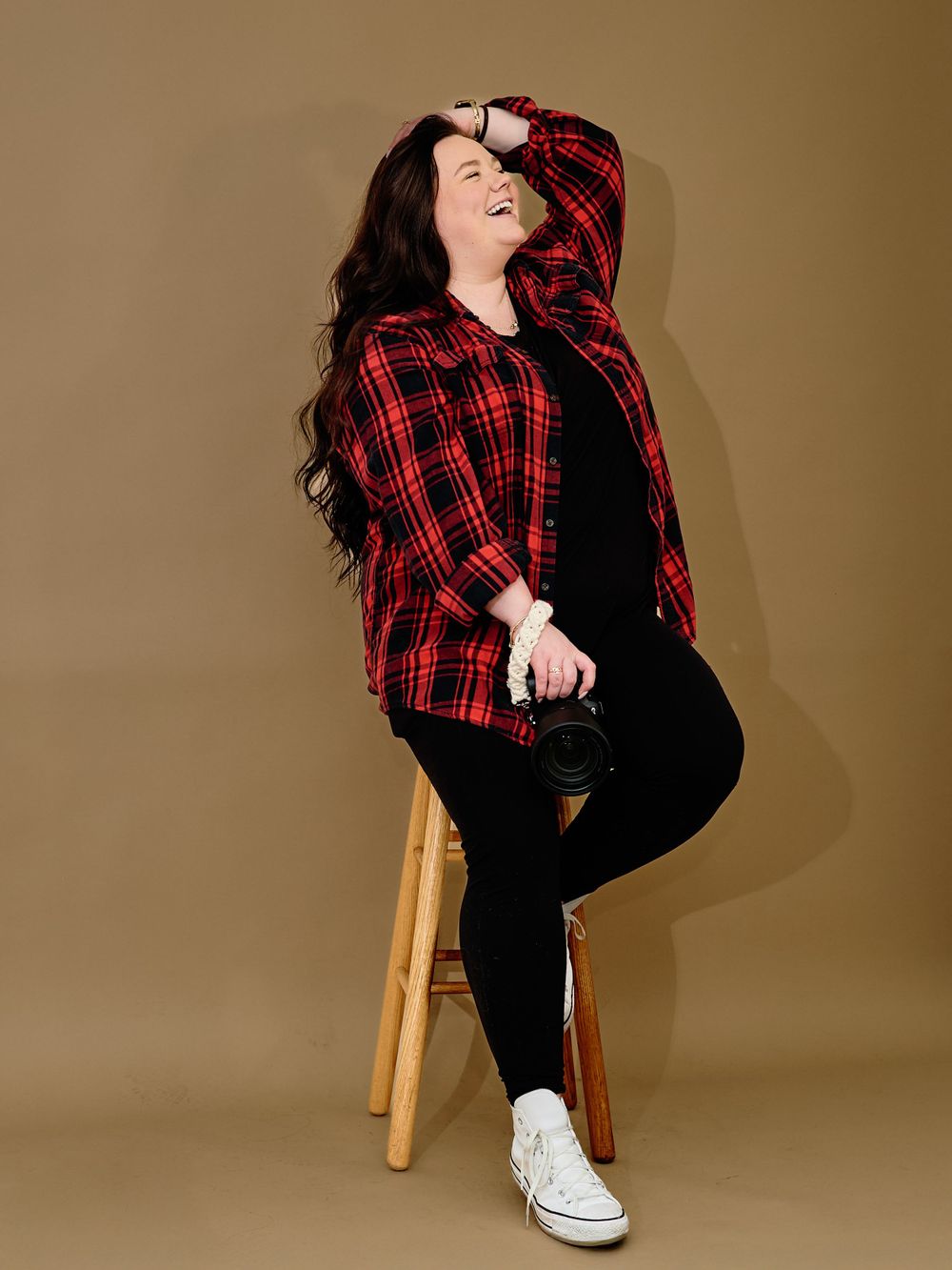 Model in plaid red flannel shirt and black pants poses casually on wooden stool against beige backdrop.