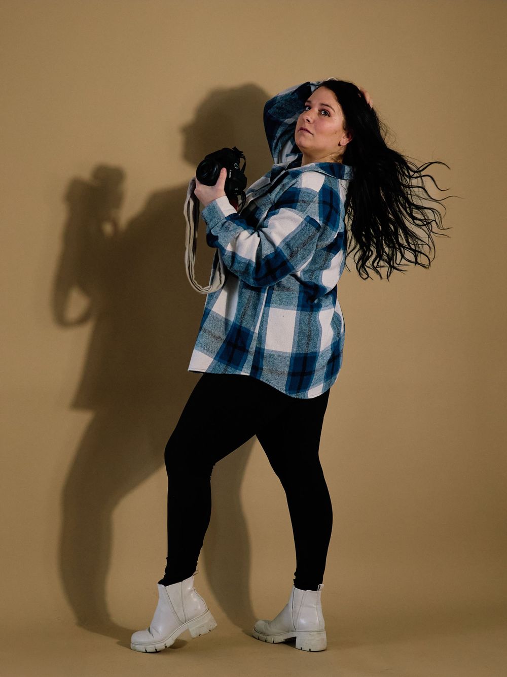 Photographer in plaid shirt and black leggings poses dynamically against beige wall with dramatic shadow effect.