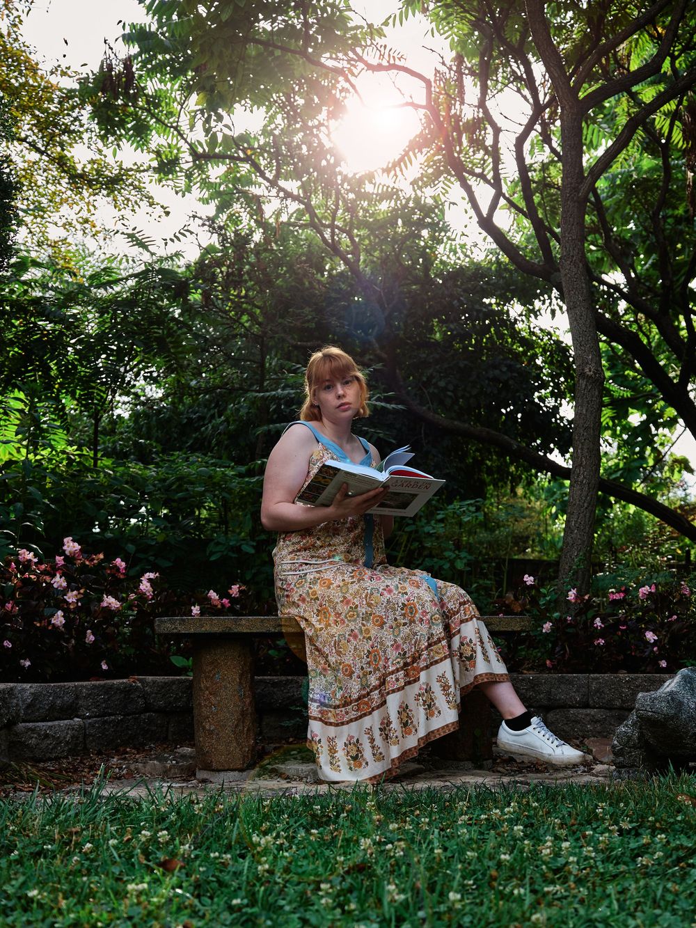 A person in a floral dress sits on a stone wall in a lush garden setting during golden hour.