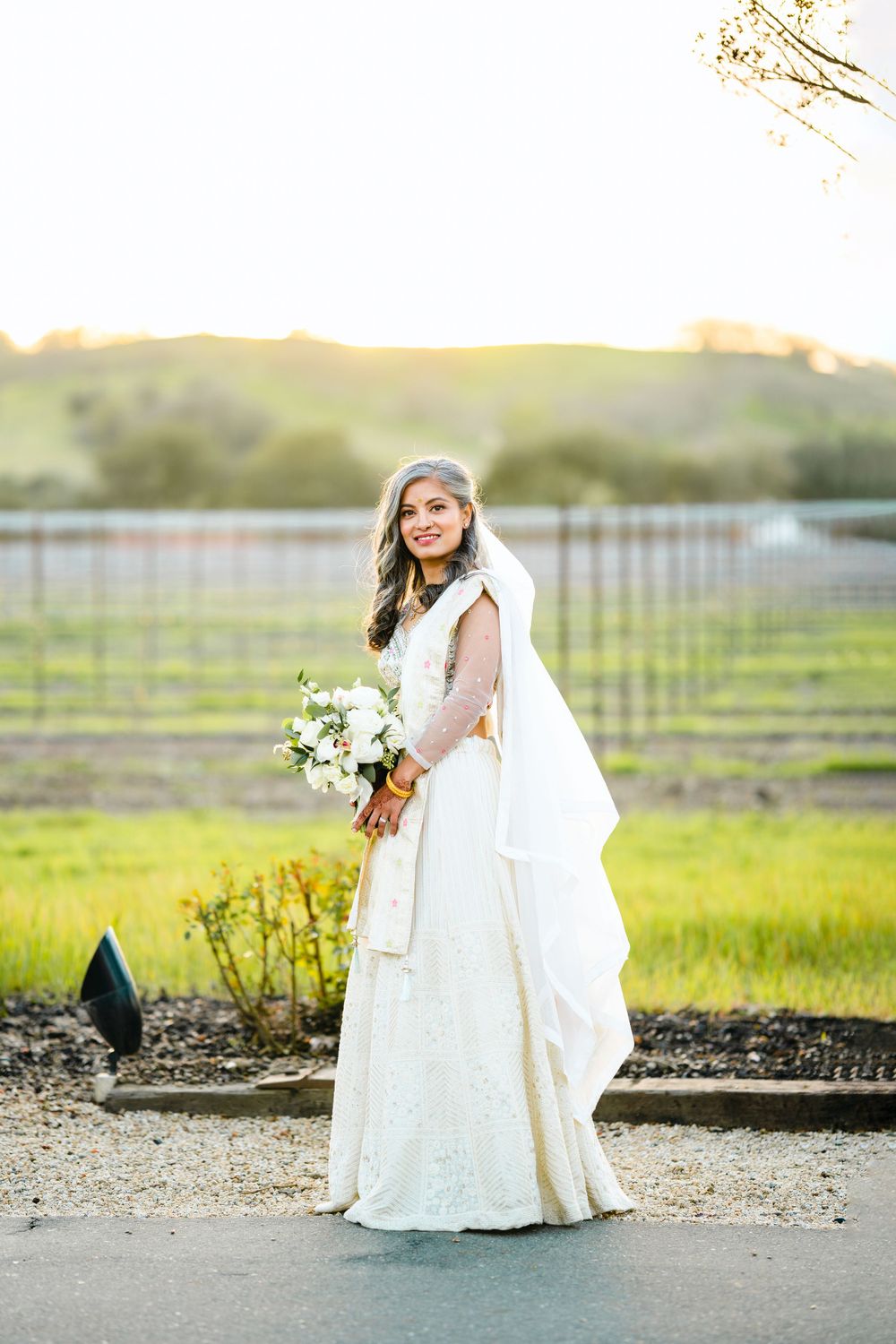 Bride in flowing white dress holds bouquet against vineyard backdrop at golden hour.