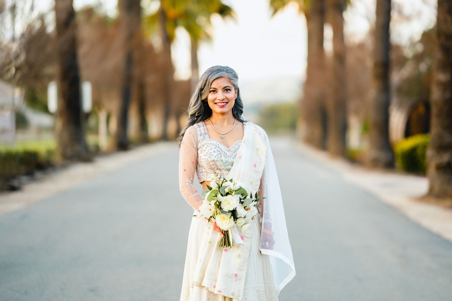 A bride in a white wedding dress poses with her bouquet on a palm tree-lined road at golden hour.