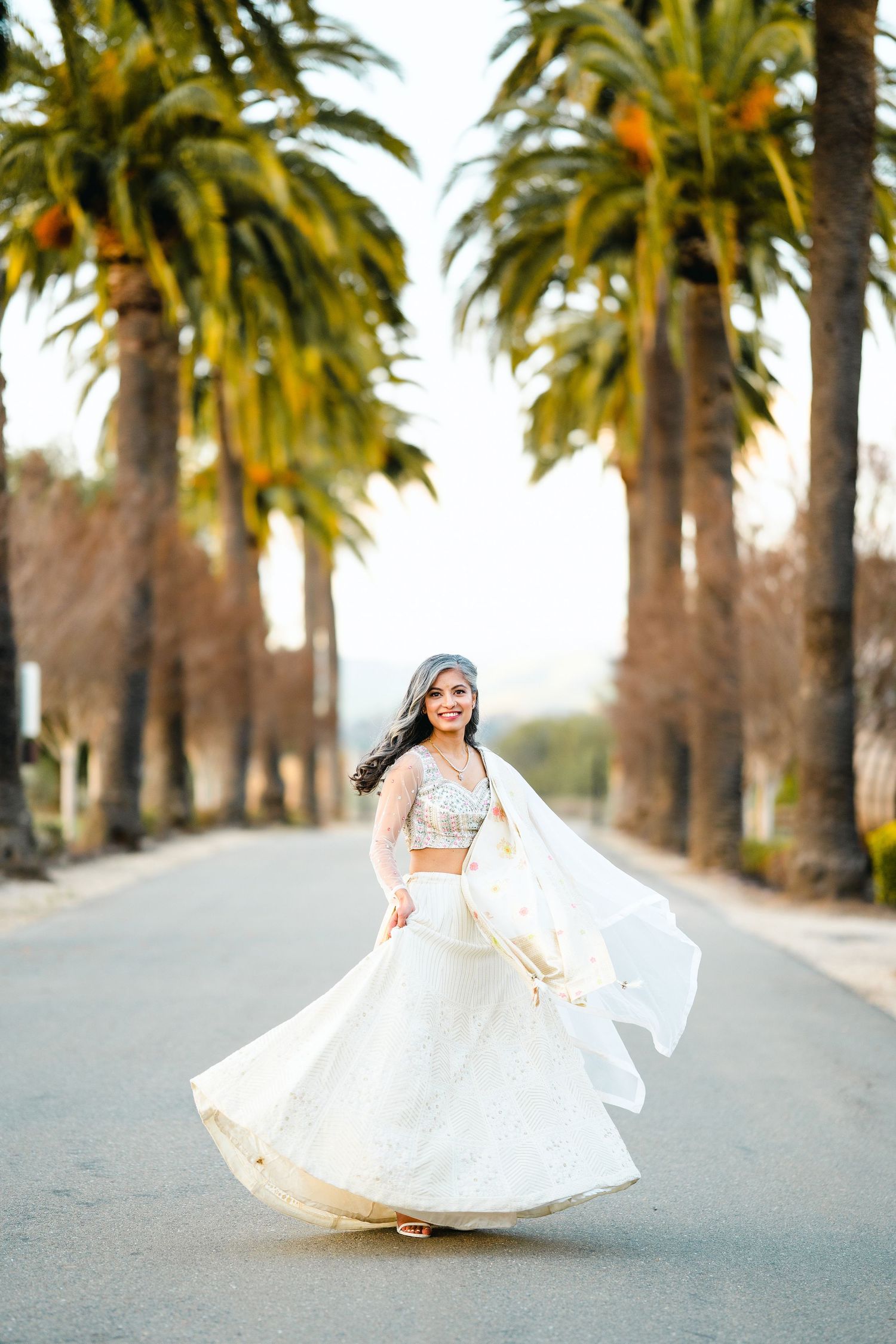 A bride twirls in her flowing white lehenga on a palm-lined street during a golden sunset.