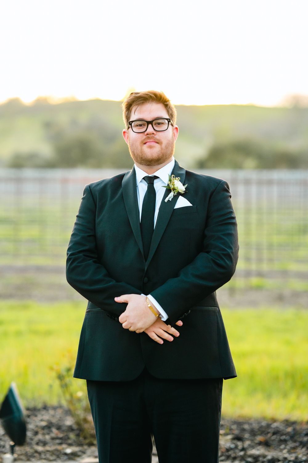 A groom in a dark suit and glasses poses in front of a vineyard at sunset.