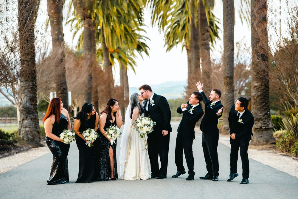 A wedding party in black attire poses together on a palm tree-lined path.
