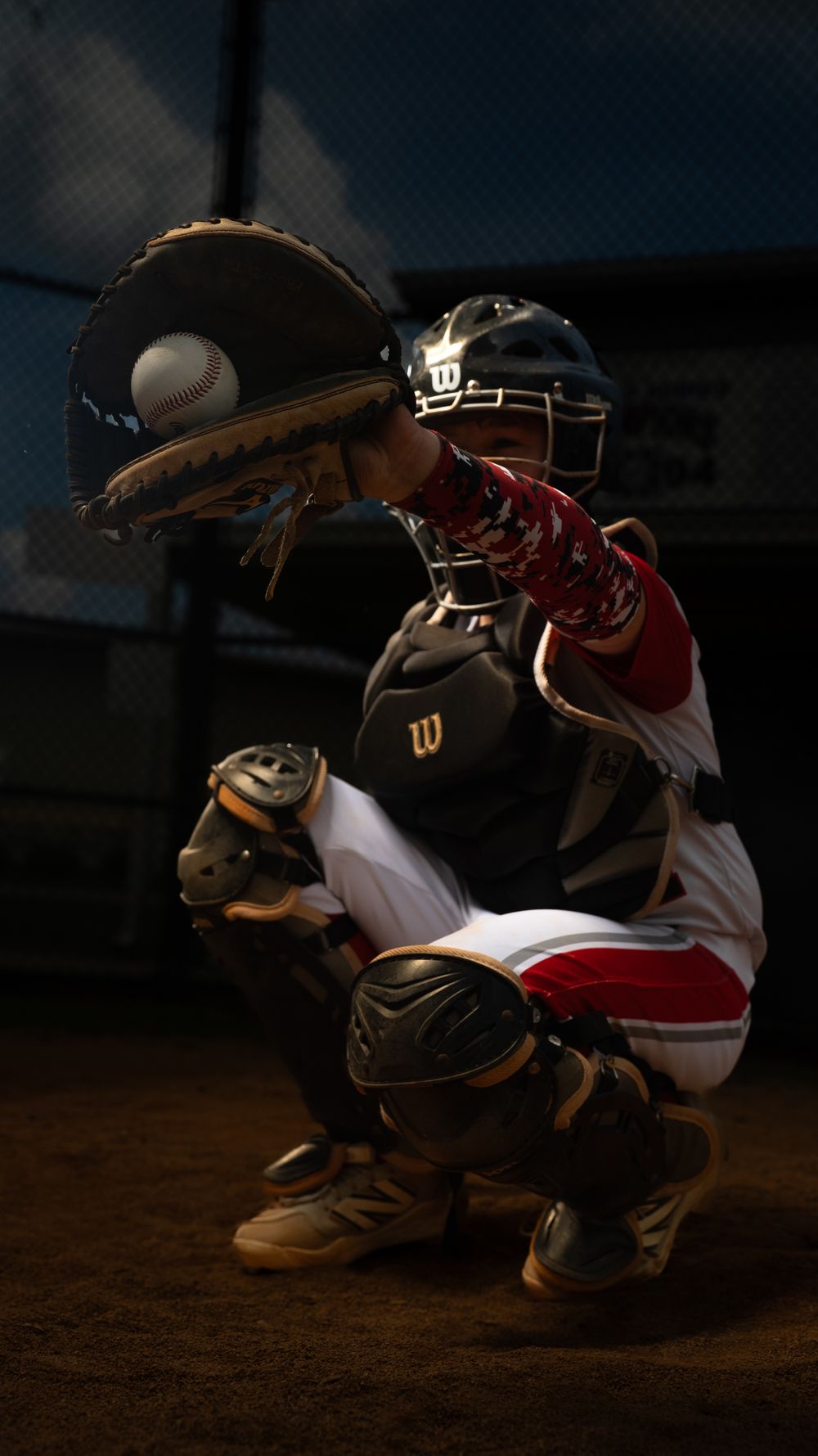Baseball catcher in protective gear crouches in position with glove ready behind home plate during night game.