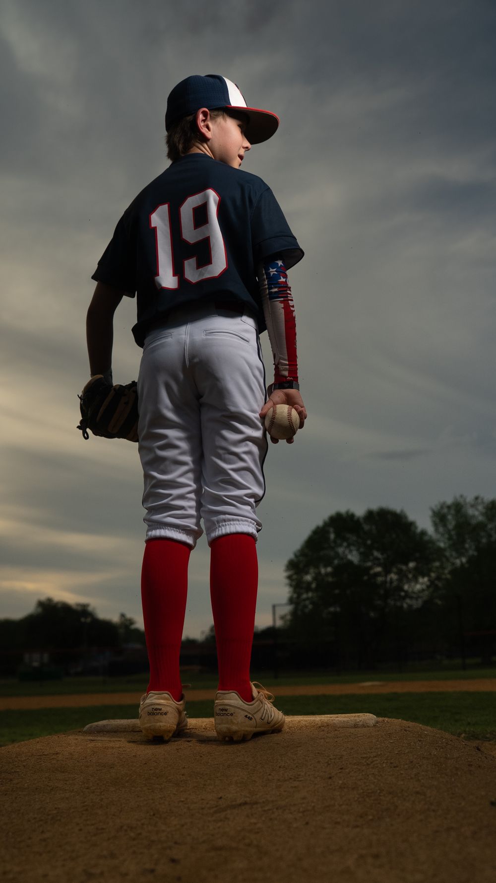 Baseball player wearing number 19 jersey stands on pitcher's mound silhouetted against dramatic cloudy sky at sunset.