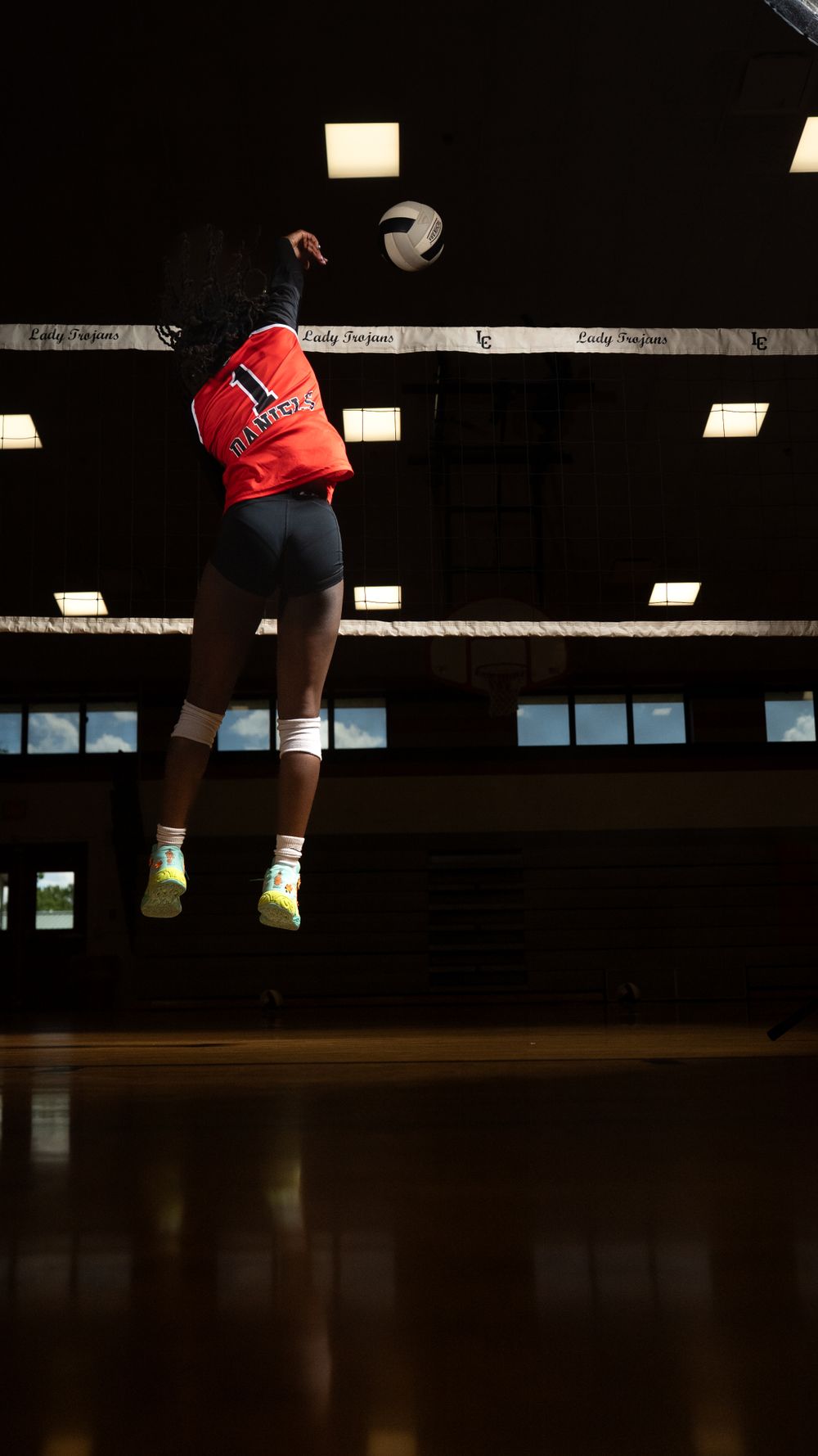 Volleyball player in red jersey leaps high to spike ball during indoor match in dark gymnasium.