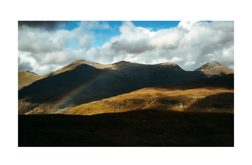 A rainbow arches over golden highland mountains under dramatic cloudy skies in a scenic landscape.