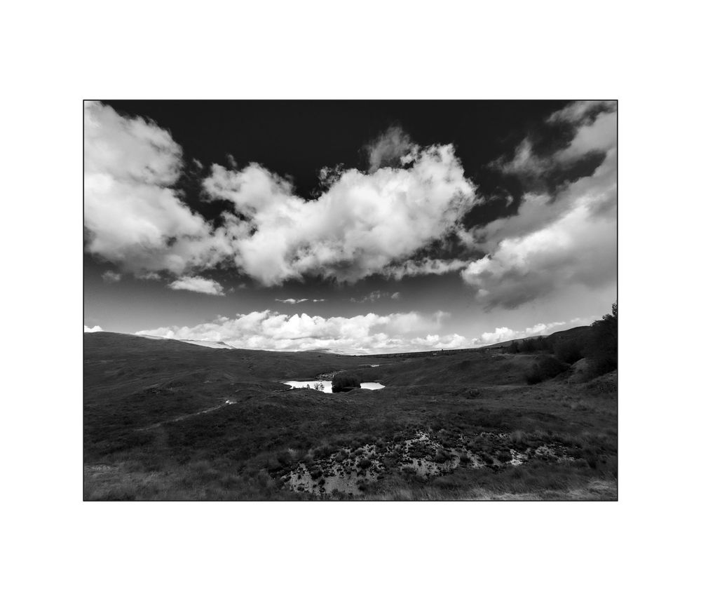 Black and white landscape photo of rolling hills under dramatic cloudy sky with a small white building visible in the valley.