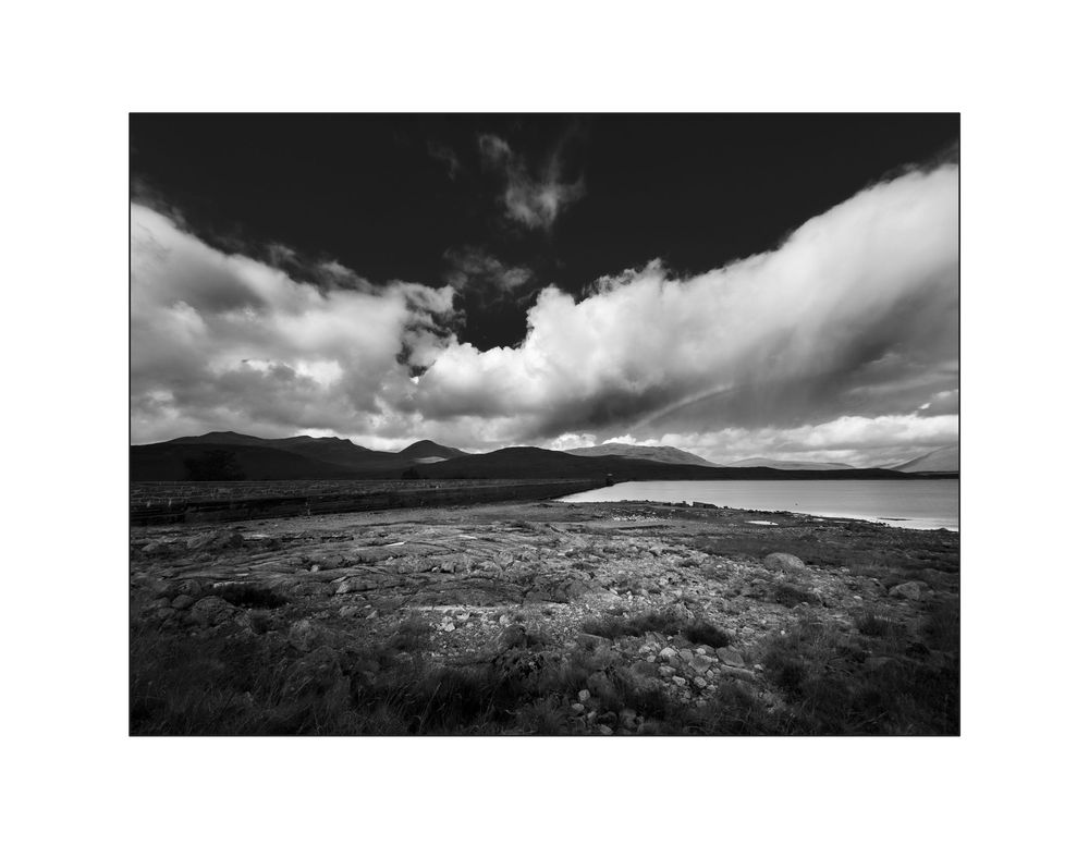 Black and white landscape photo of a rugged moorland with a loch, mountains, and dramatic cloudy sky.