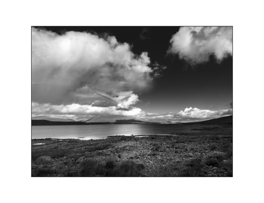 Black and white landscape photo of a calm loch under dramatic cloudy skies with rolling hills in the distance.
